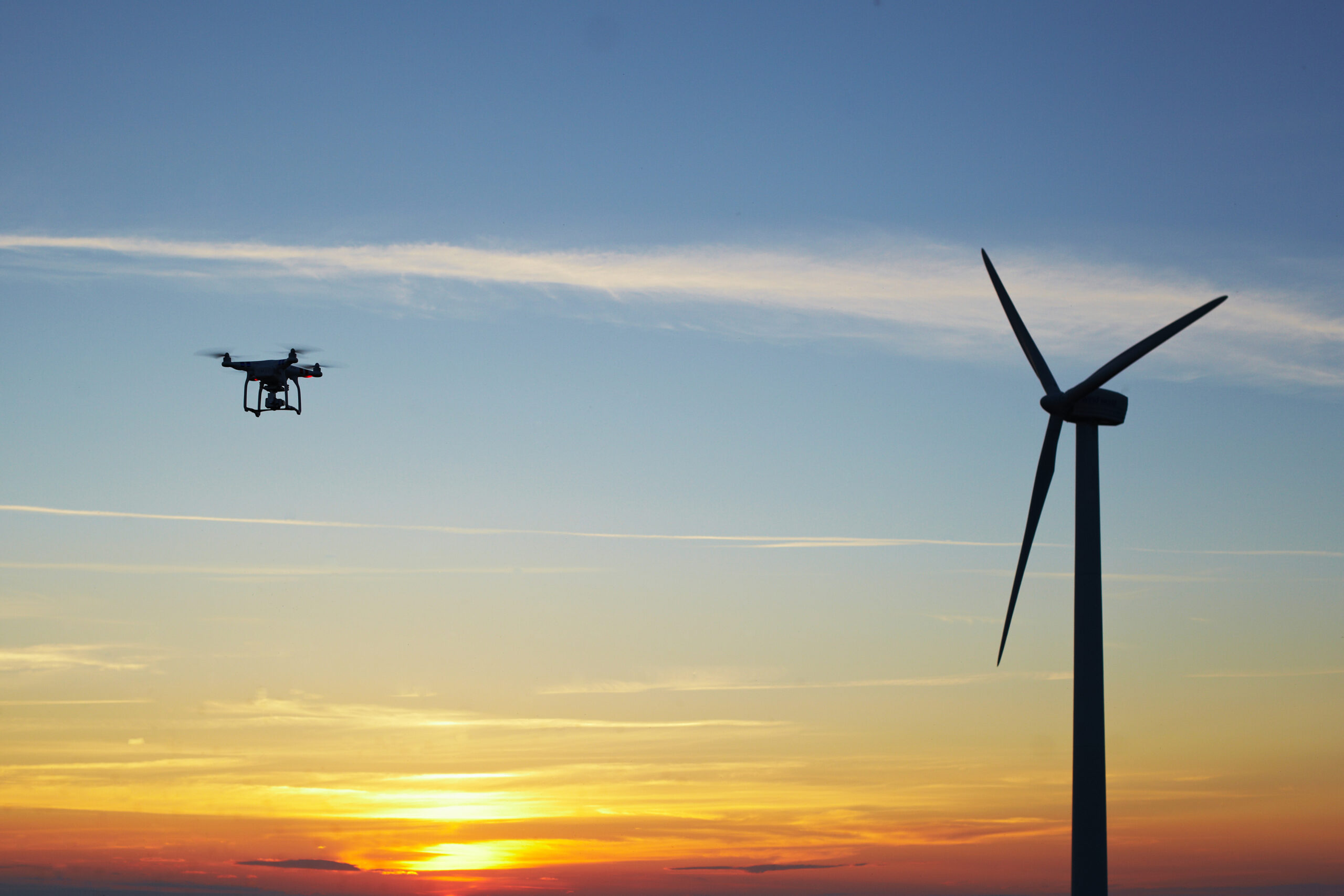 A photo of a drone hovering near a wind turbine at sunset.