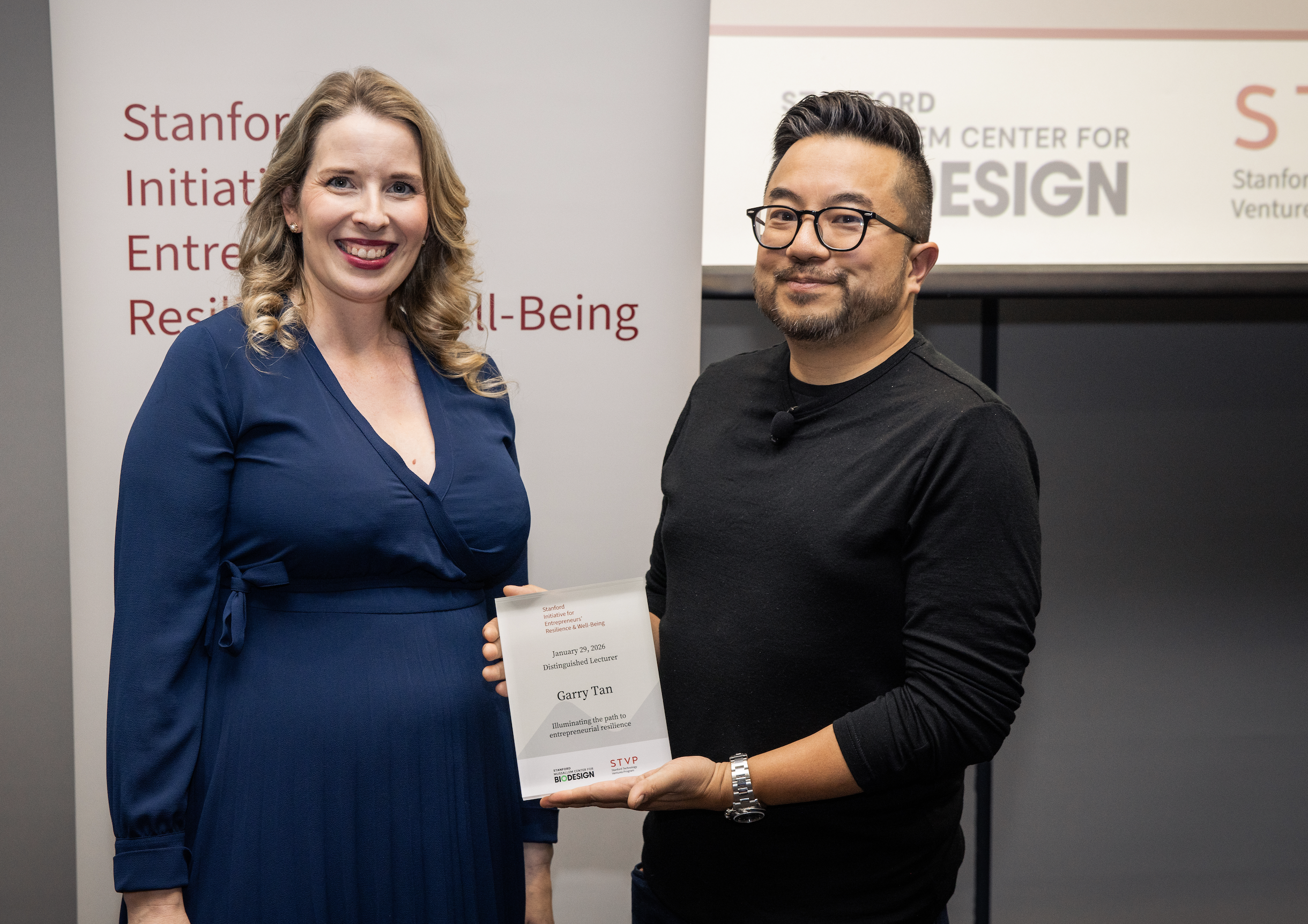 Y Combinator CEO Garry Tan and STVP Executive Director Ashlea Wallington stand together for a picture. Ashlea is wearing a blue dress, Garry is wearing a black shirt and holding a plaque.